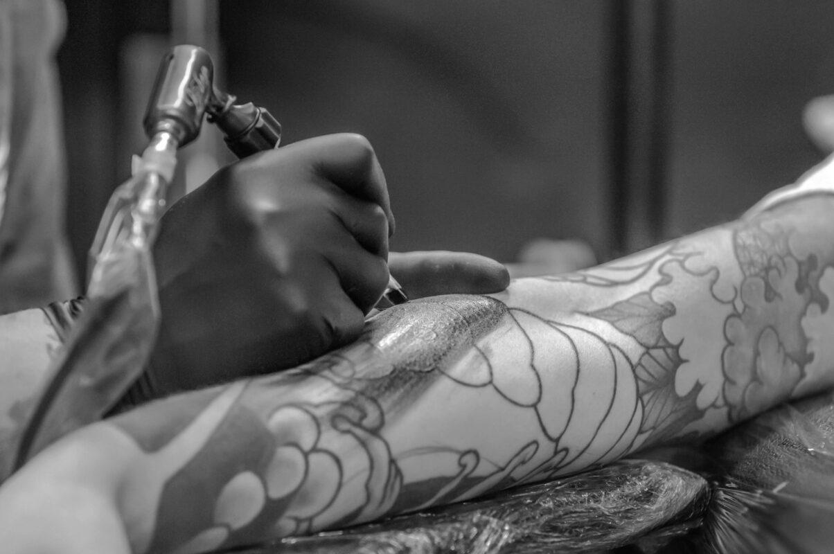 Black and white close-up of a tattoo machine shading a large floral design on a client's arm